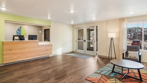 A reception area with a wooden desk, a chair, a floor lamp, and a view of the outside through a glass door. - WoodSpring Suites Thornton-North Denver (Thornton)