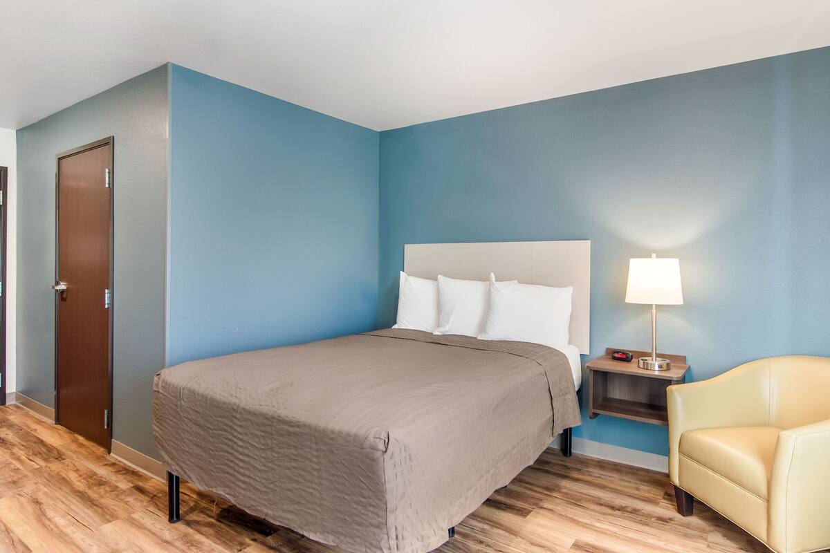 Ein Hotelzimmer mit einem Bett, einem Nachttisch mit Lampe, einem Sessel und einer Tür.