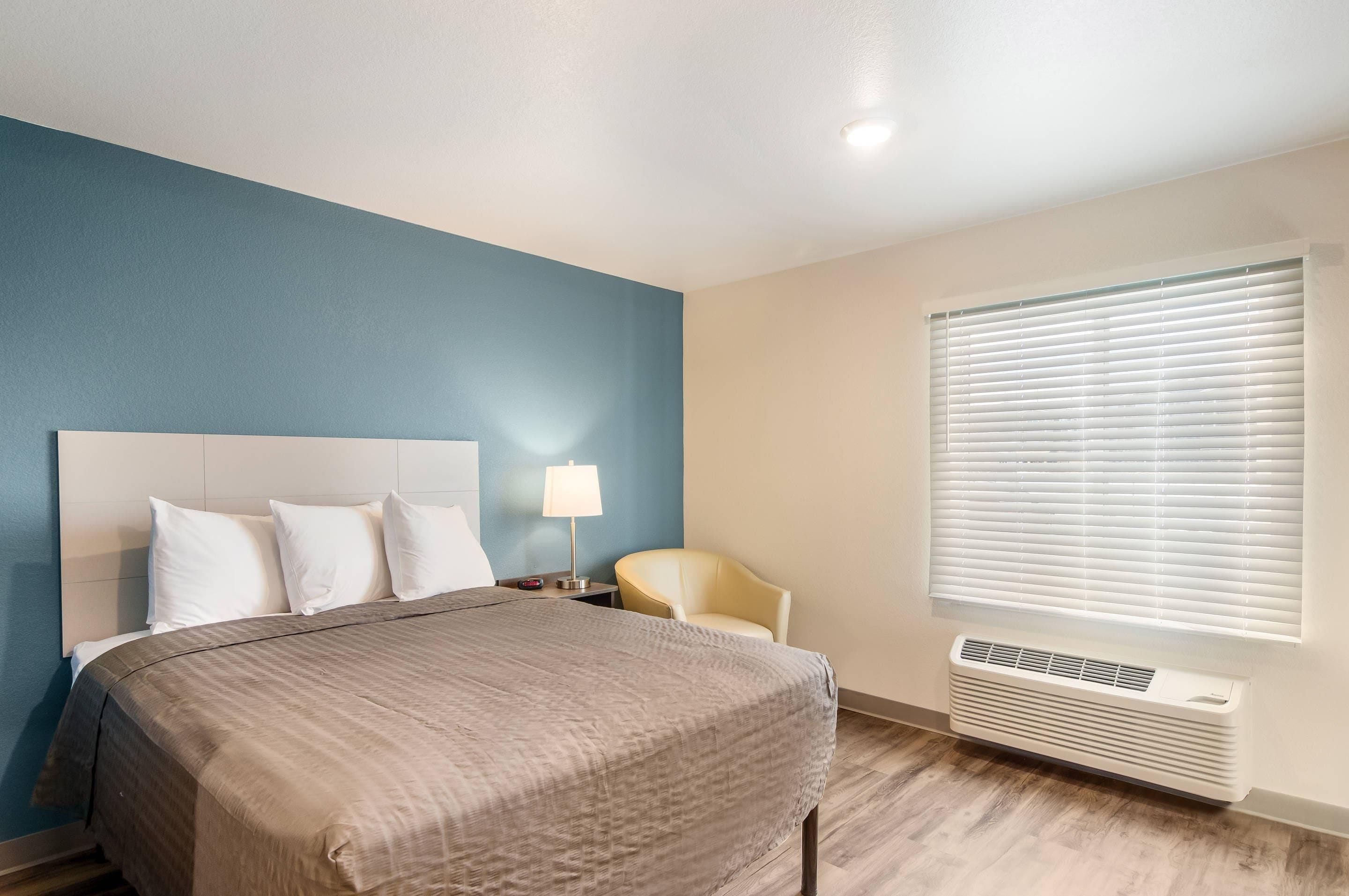 Habitación de hotel con cama, una silla, mesita de noche con lámpara, ventana con persianas y aire acondicionado instalado en la pared.