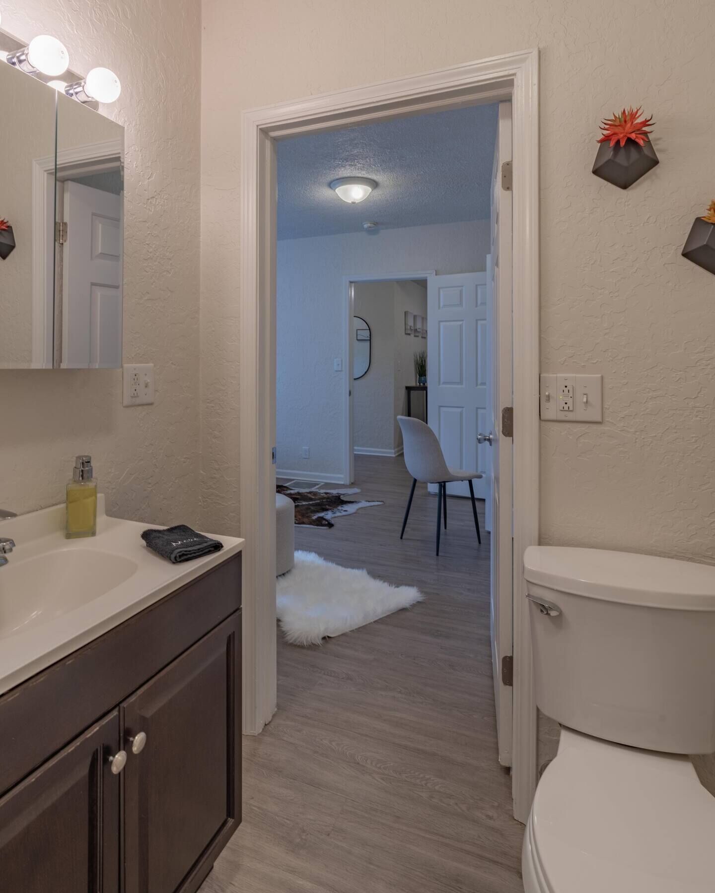 Un baño con lavamanos blanco, muebles de madera oscura, inodoro blanco y espejo con iluminación.