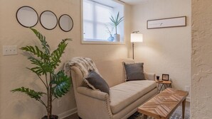 A cozy living room with a beige sofa, a wooden coffee table, a potted plant, and a window with blinds. - University Suite "C" (Marion)