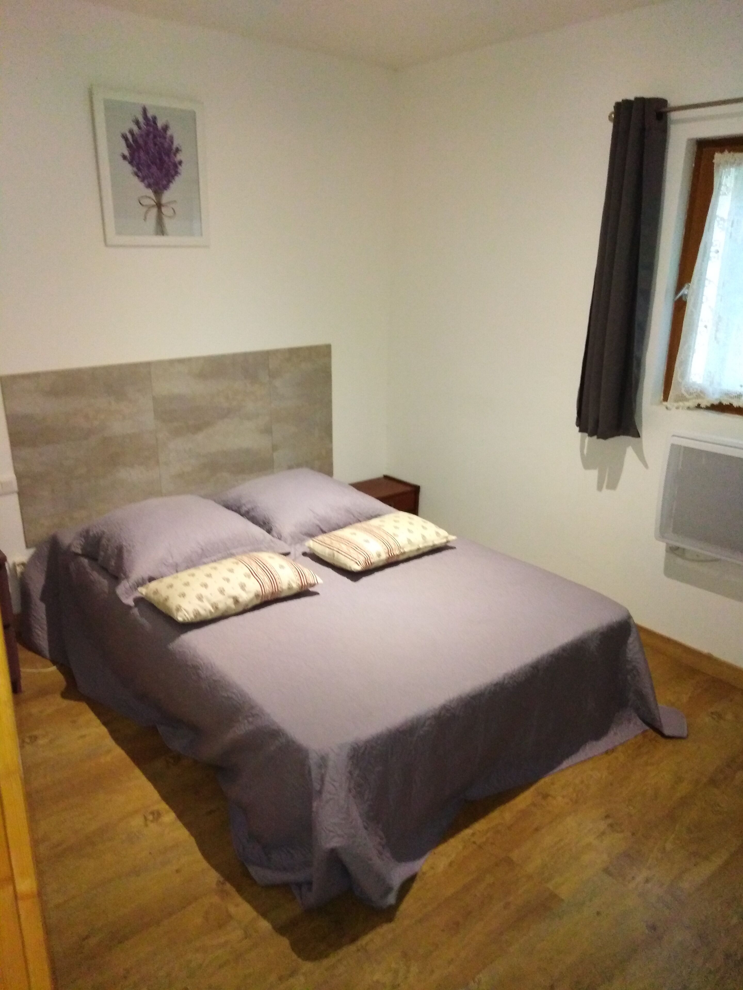 A bedroom with a bed, pillows, a nightstand, a window with curtains, and a wall-mounted picture.