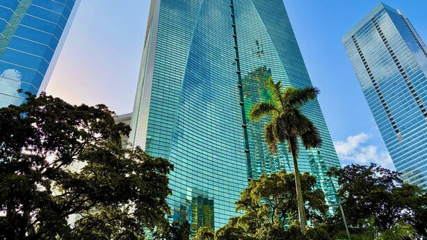 Un grand bâtiment moderne en verre, doté d’une façade réfléchissante, entouré d’arbres et d’autres immeubles.