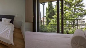 Une chambre dotée d’un grand lit, d’un plancher en bois et d’un balcon offrant une vue sur la verdure et les bâtiments.