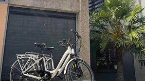 Un bâtiment moderne à deux étages, doté d’une porte de garage, avec un vélo blanc garé devant et un palmier à proximité.