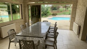 Un patio couvert avec une longue table à manger et des chaises, une vue sur une piscine et des murs en pierre.