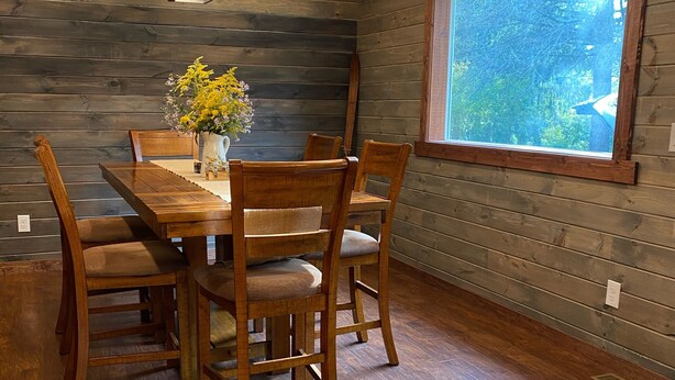 A dining area with a wooden table and chairs, a vase with flowers, and a window with a view of trees.