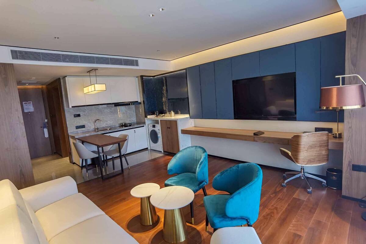 Un salon moderne avec un canapé, deux tables rondes et des chaises bleues. On aperçoit, au fond, une cuisine avec une table à manger et des chaises.