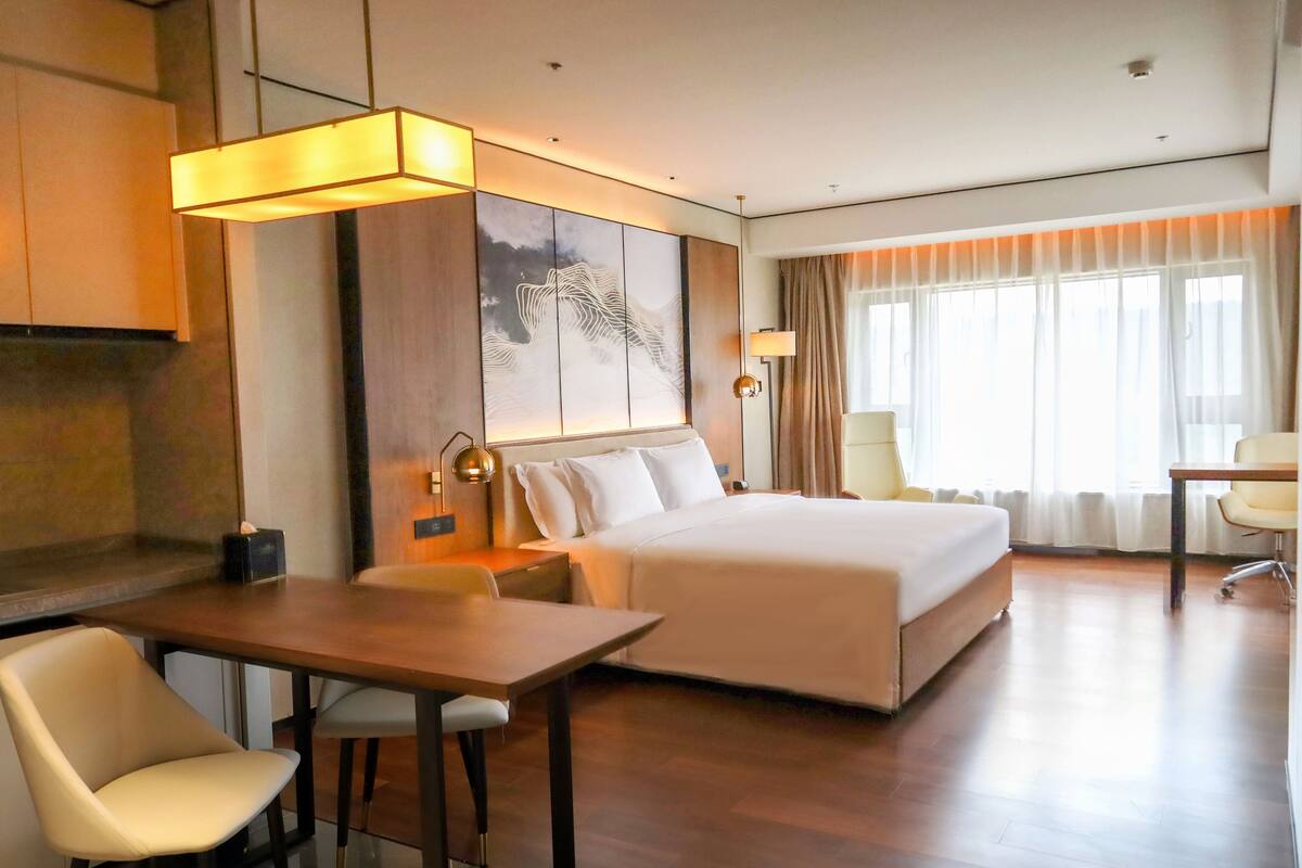 Une chambre d’hôtel avec un grand lit, un bureau et une chaise. Il y a un tableau au mur et une fenêtre avec des rideaux.