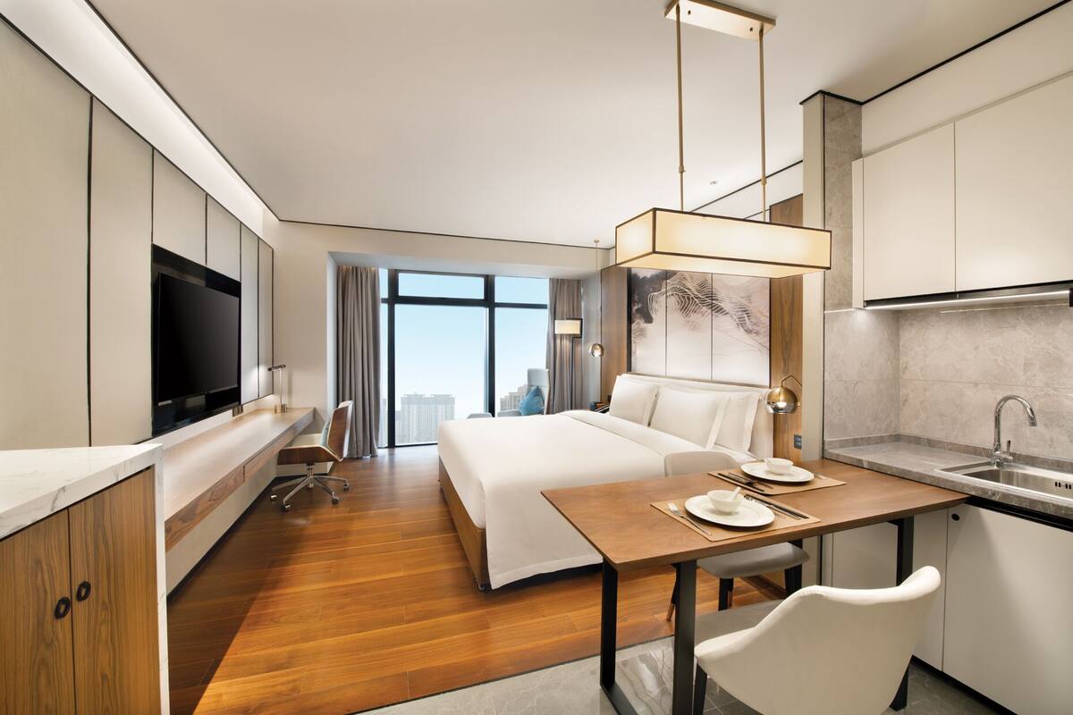 Une chambre d’hôtel moderne dotée d’un grand lit, d’un bureau, d’une télévision à écran plat, d’une table à manger avec des chaises et d’un coin cuisine.