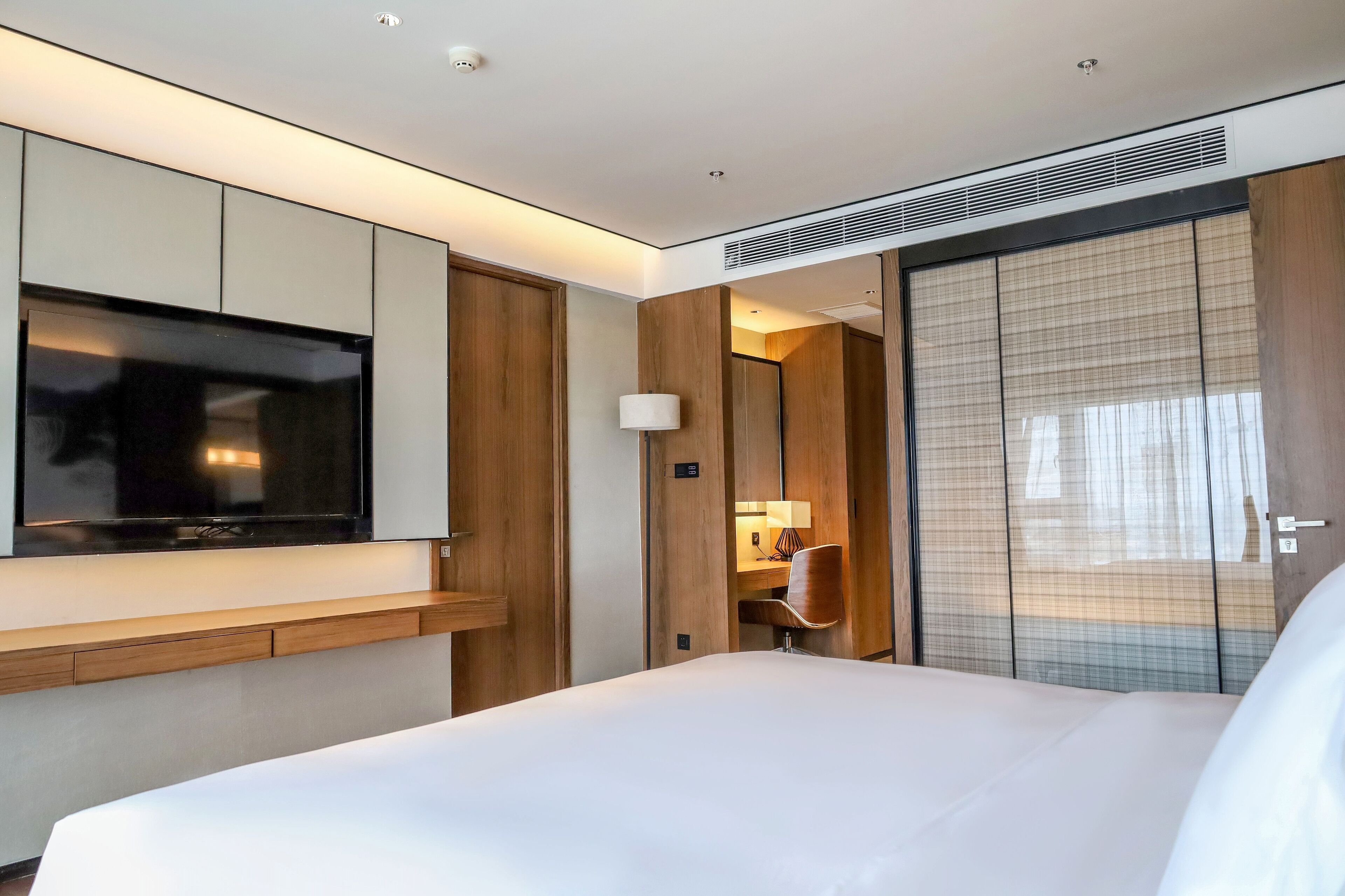 A modern hotel room with a large bed, a flat-screen TV mounted on the wall, a wooden desk with a chair, and a sliding door with a view.