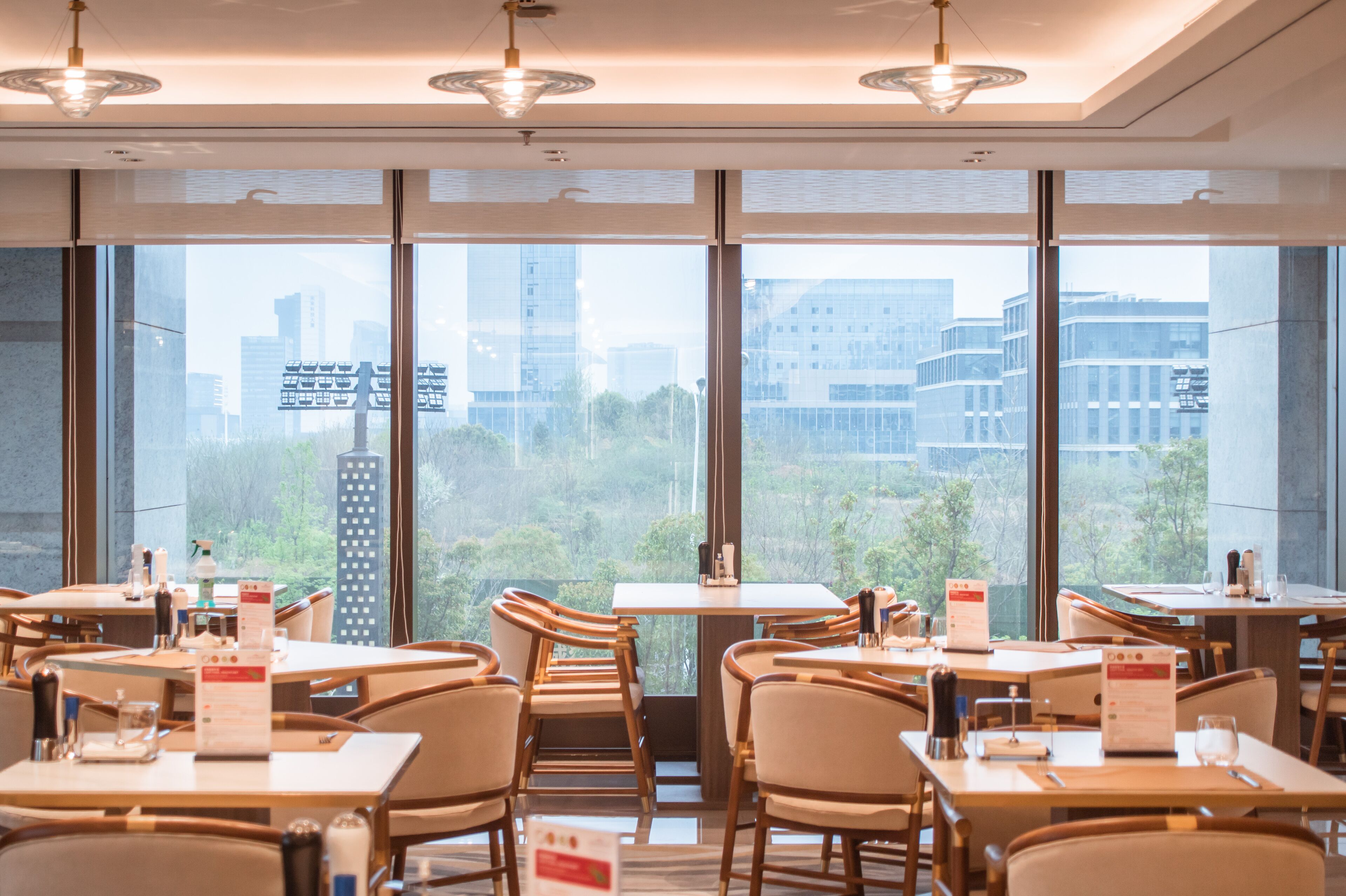 A modern dining area with tables and chairs, large windows with a city view, and pendant lighting.