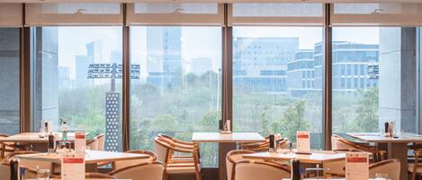 A modern dining area with tables and chairs, large windows with a city view, and pendant lighting.
