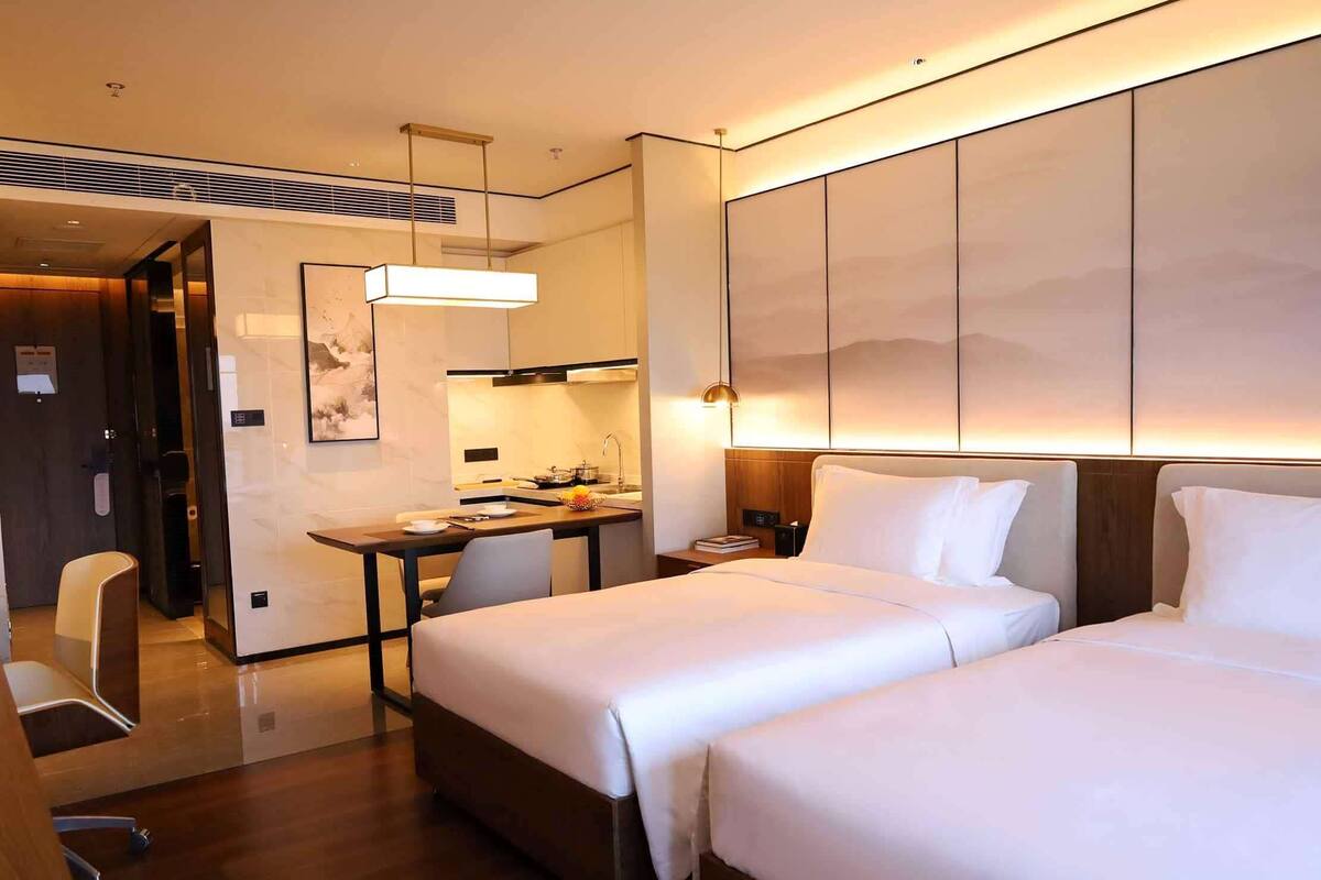 Une chambre d’hôtel avec deux lits, un bureau, une chaise, un petit coin cuisine et un grand mur donnant sur une vue sur la montagne.