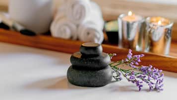 Un ambiente de spa con una cuenca de piedra, toallas enrolladas, una planta en maceta, velas y una rama de lavanda.