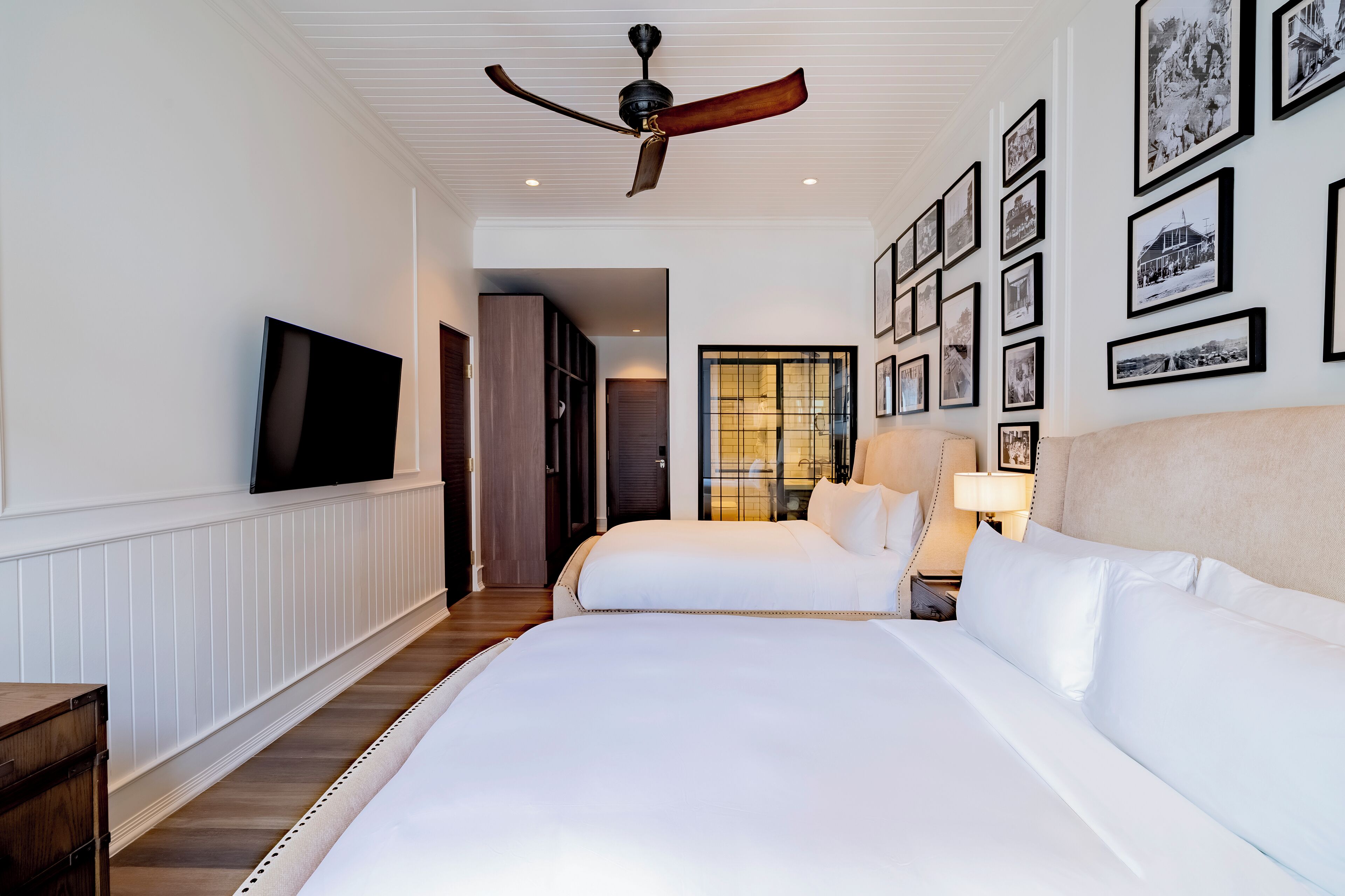 A hotel room with two beds, a ceiling fan, a TV, and framed pictures on the wall.