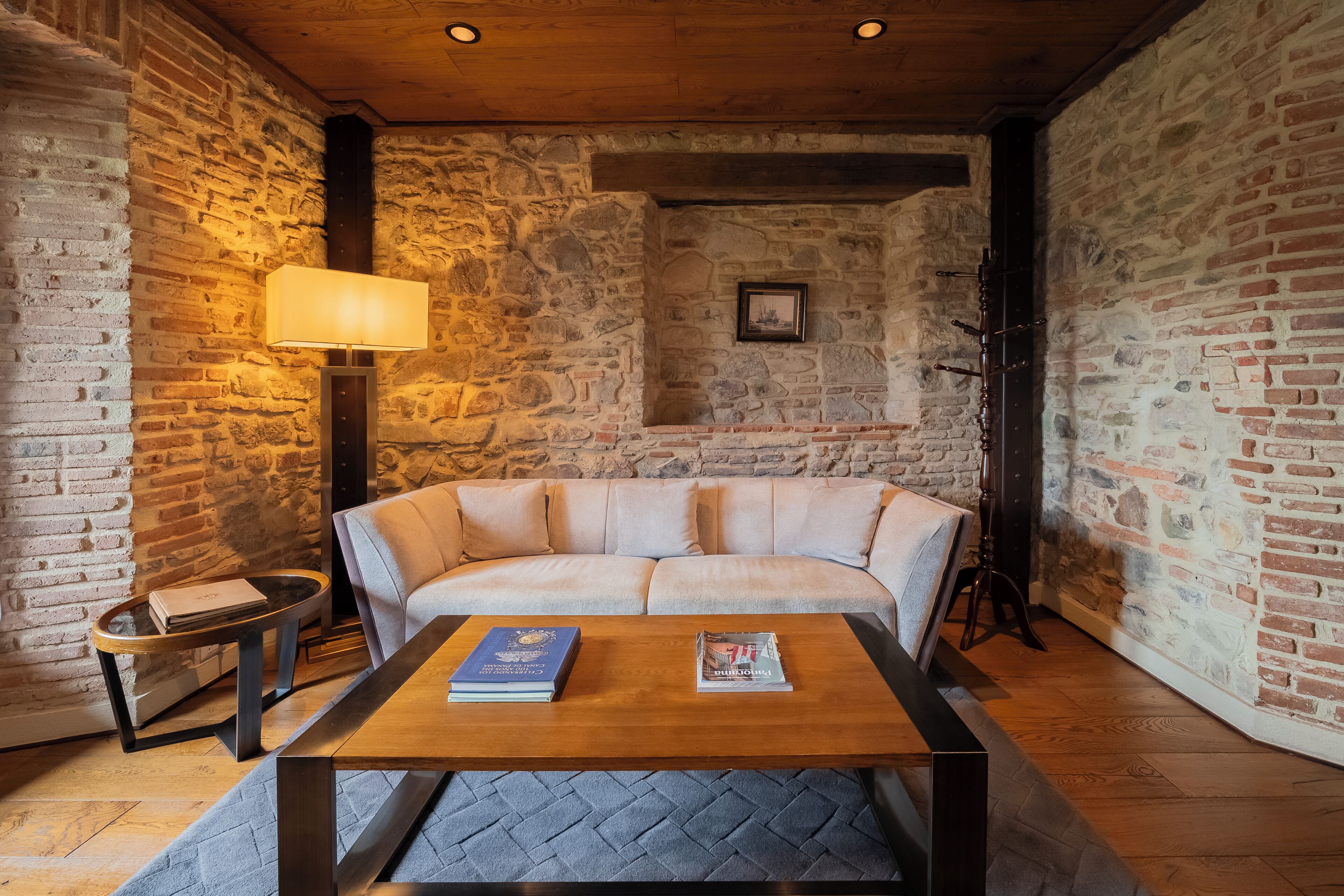 A cozy room with a stone wall, a wooden ceiling, a cushioned sofa, a coffee table with books, a lamp, and a framed picture on the wall.