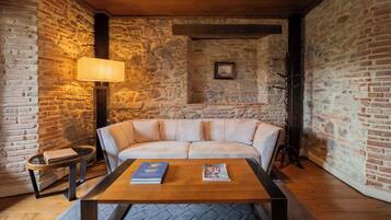 A cozy room with a stone wall, a wooden ceiling, a cushioned sofa, a coffee table with books, a lamp, and a framed picture on the wall.