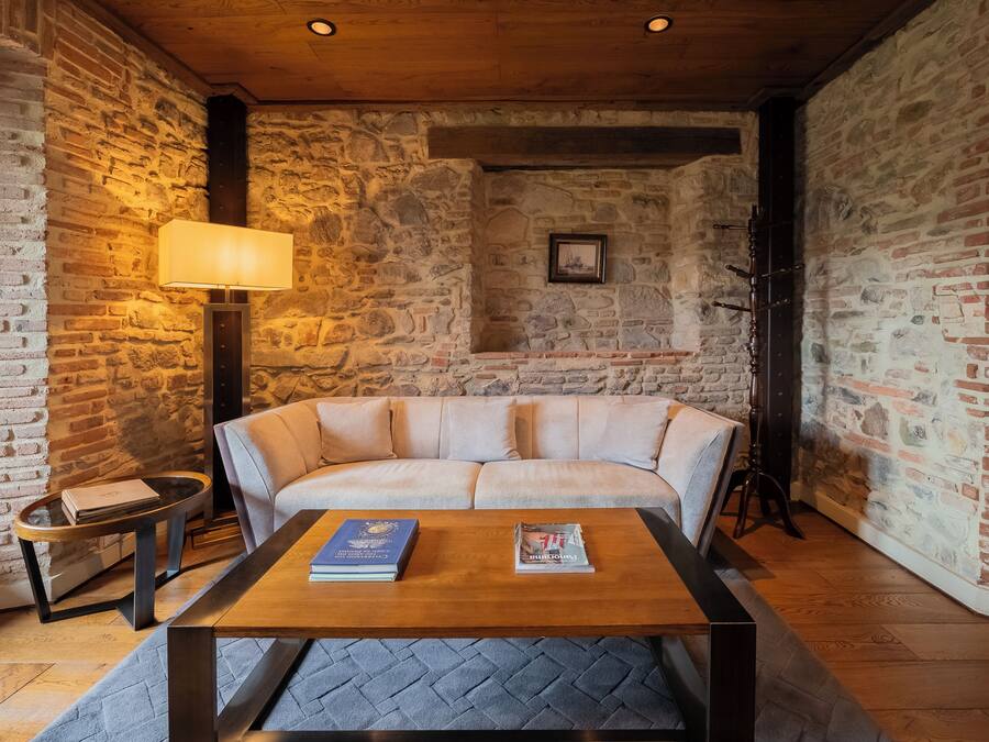 A cozy room with a stone wall, a wooden ceiling, a cushioned sofa, a coffee table with books, a lamp, and a framed picture on the wall.
