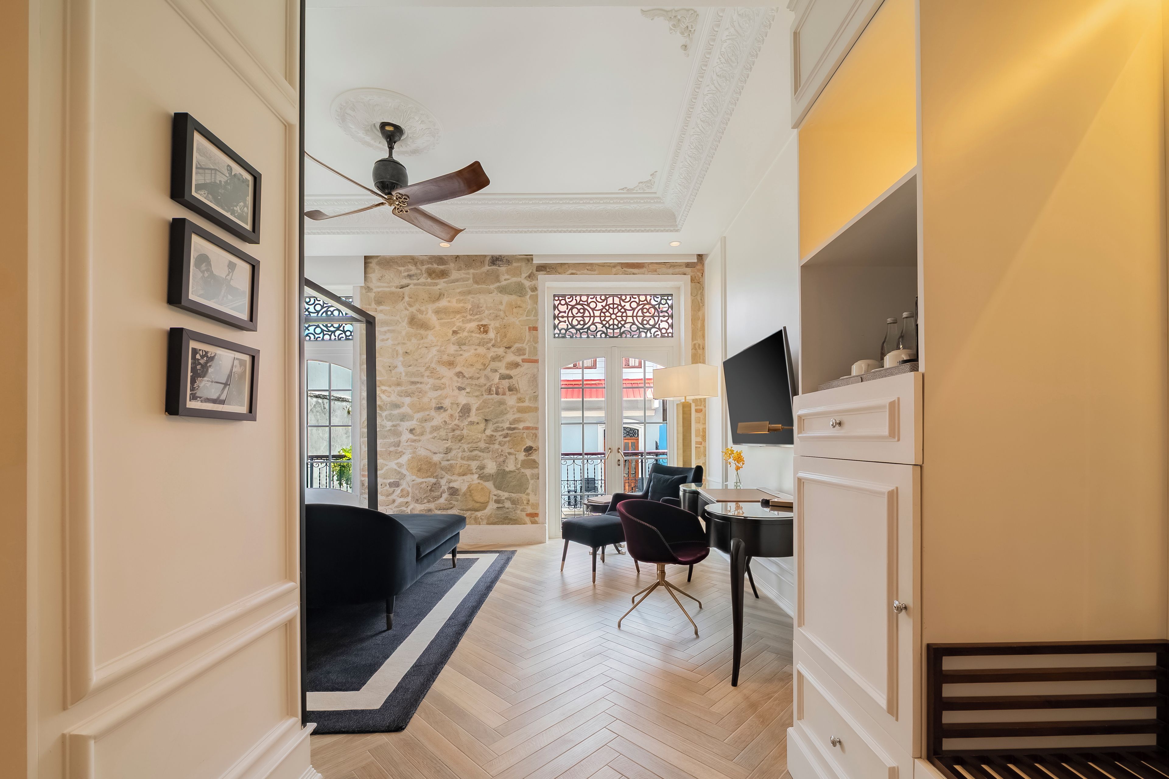 A hallway with a stone wall, a ceiling fan, a black sofa, a dining table with chairs, and a TV mounted on the wall.