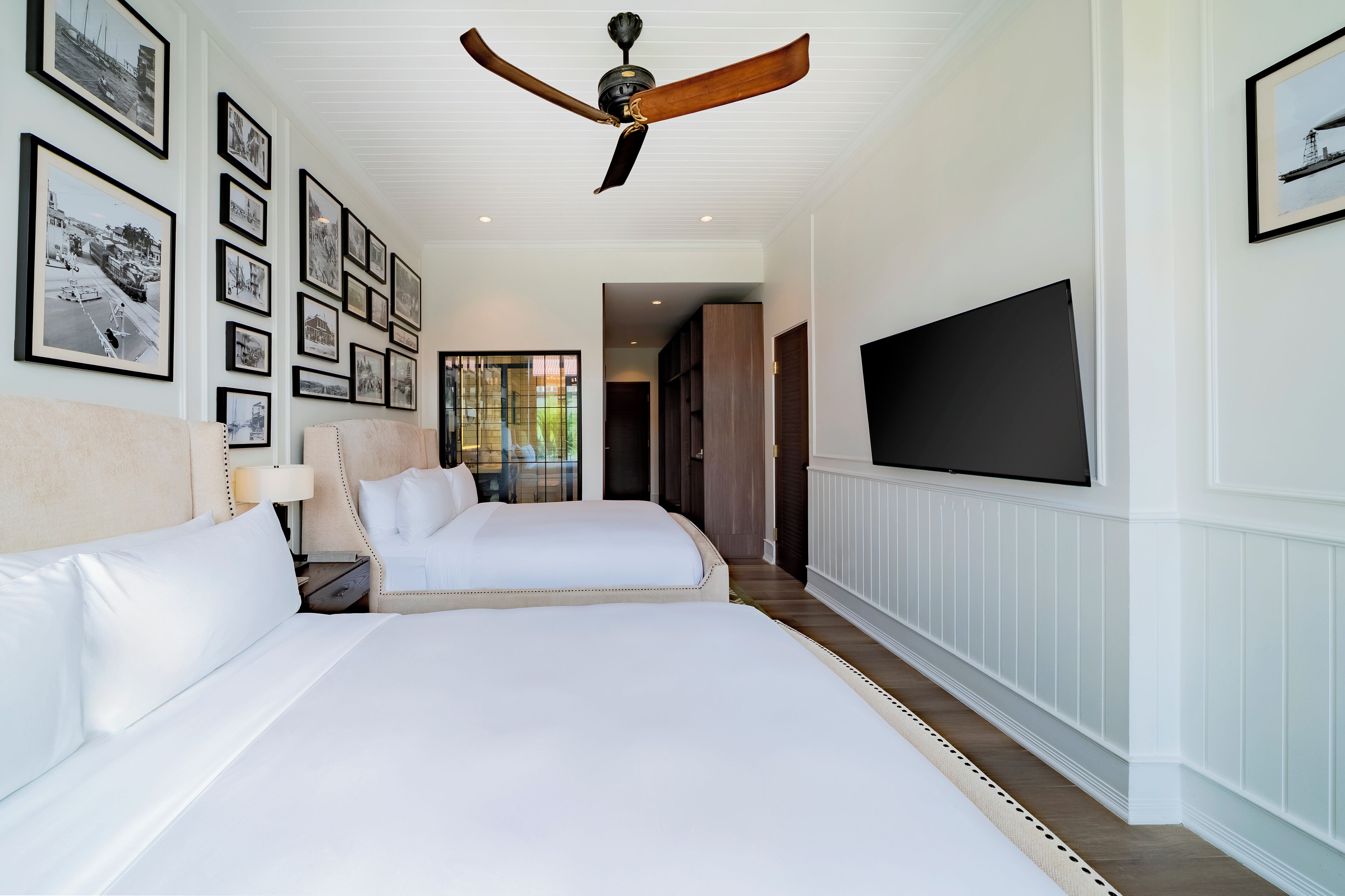 A hotel room with two beds, a ceiling fan, a TV, and framed pictures on the wall.