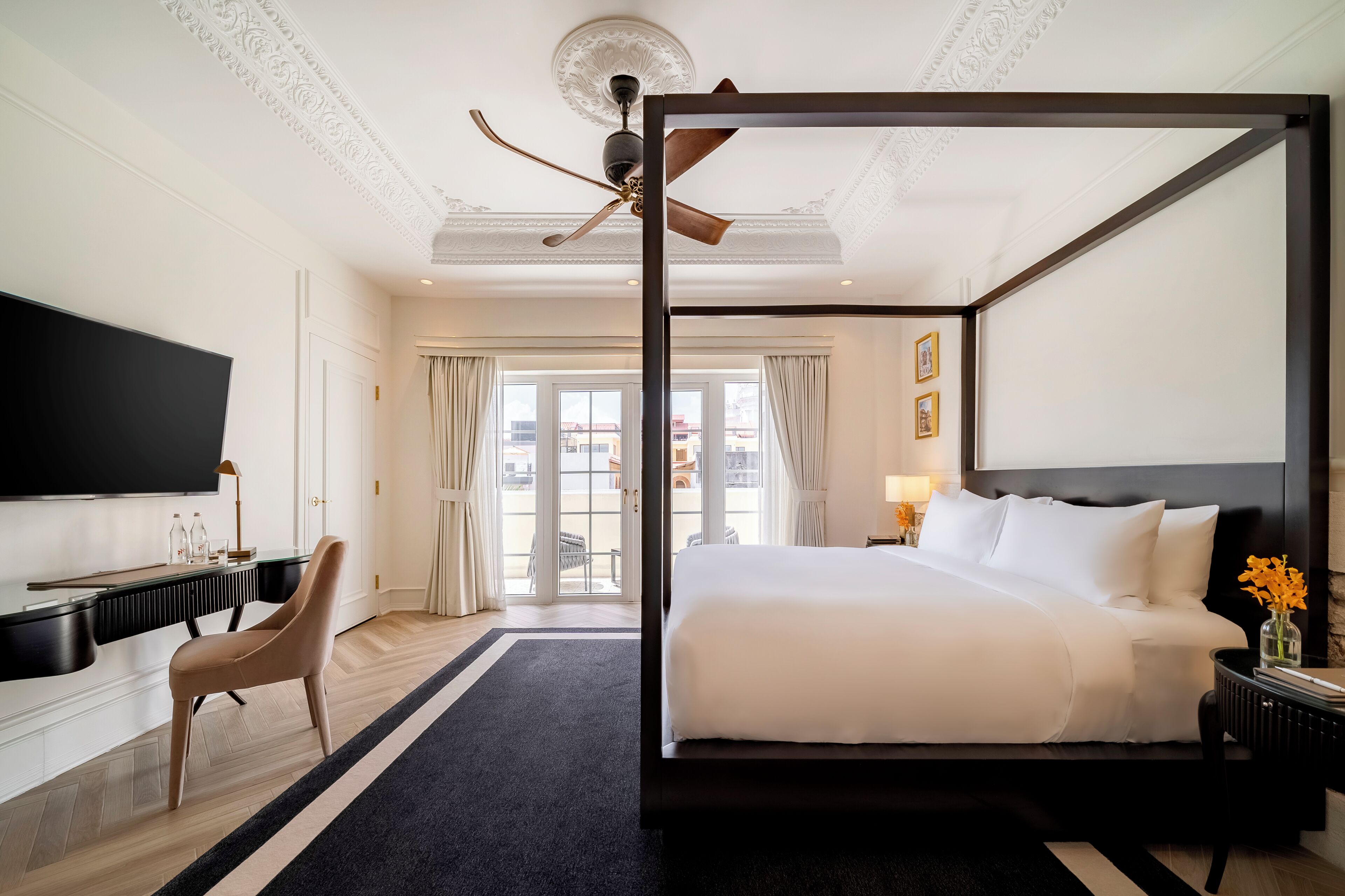A spacious bedroom with a four-poster bed, a flat-screen TV, a desk with a chair, and a large window with curtains.