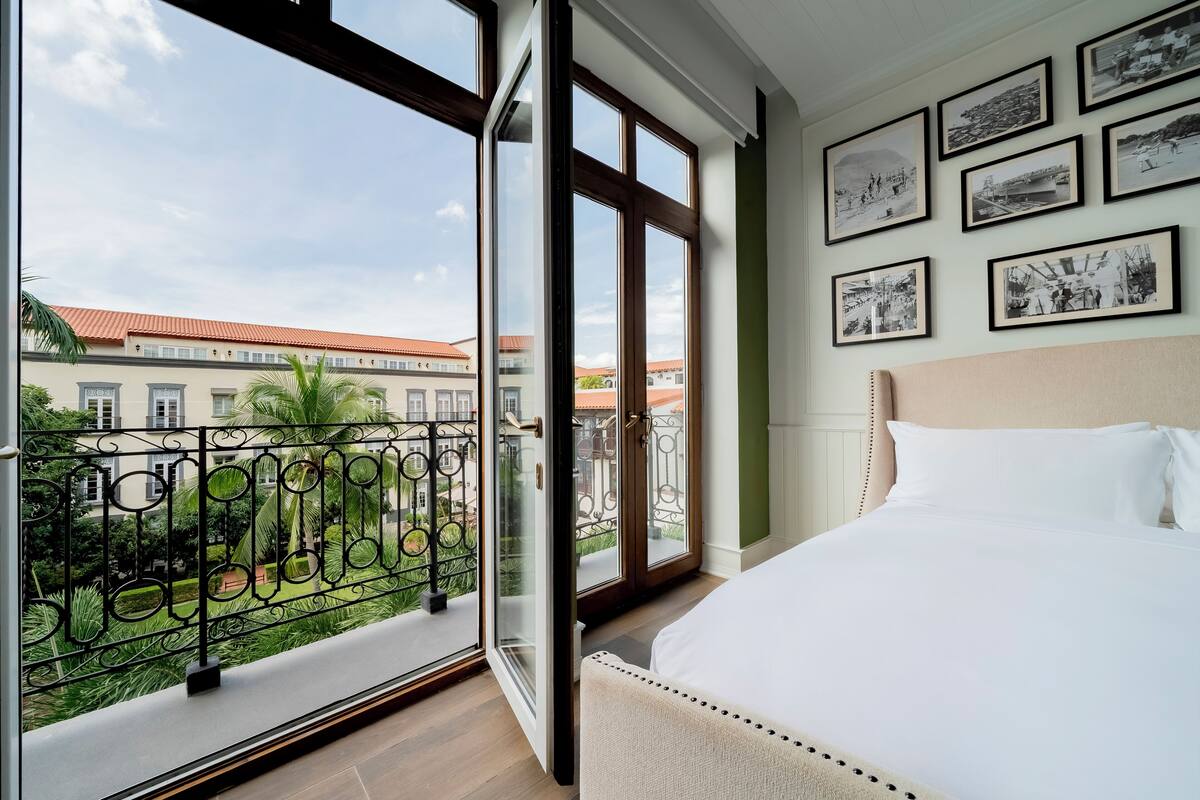 A hotel room with a large bed, a balcony with a wrought iron railing, and a view of buildings and greenery.