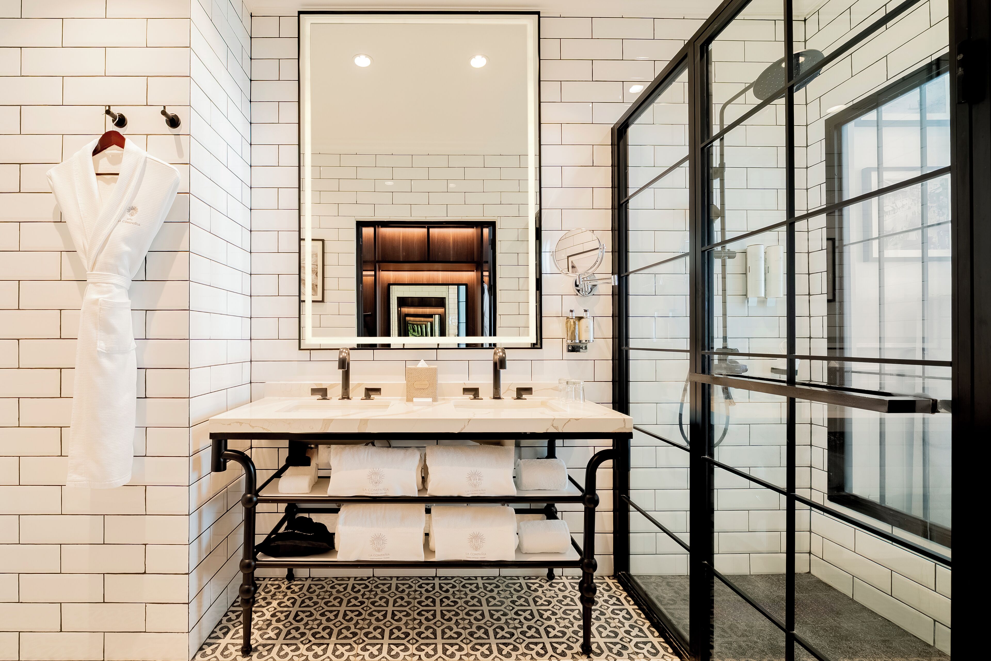 A modern bathroom with white tiled walls, a large mirror, a double sink, and a glass shower enclosure.