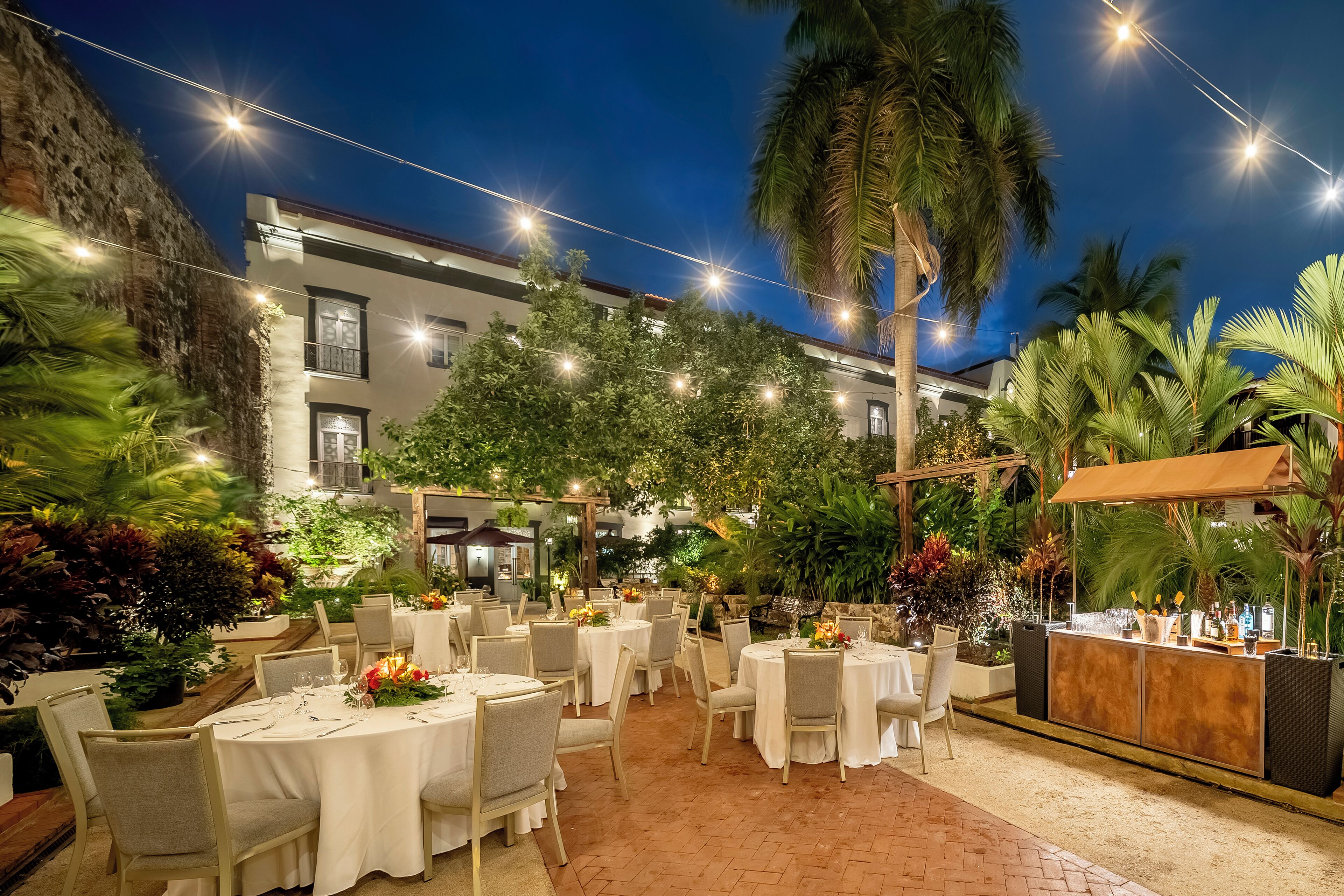 An outdoor dining area with tables set for guests, surrounded by lush greenery and palm trees.