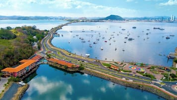 An aerial view of a coastal marina with multiple boats, a curved road, and a hotel with a red roof.