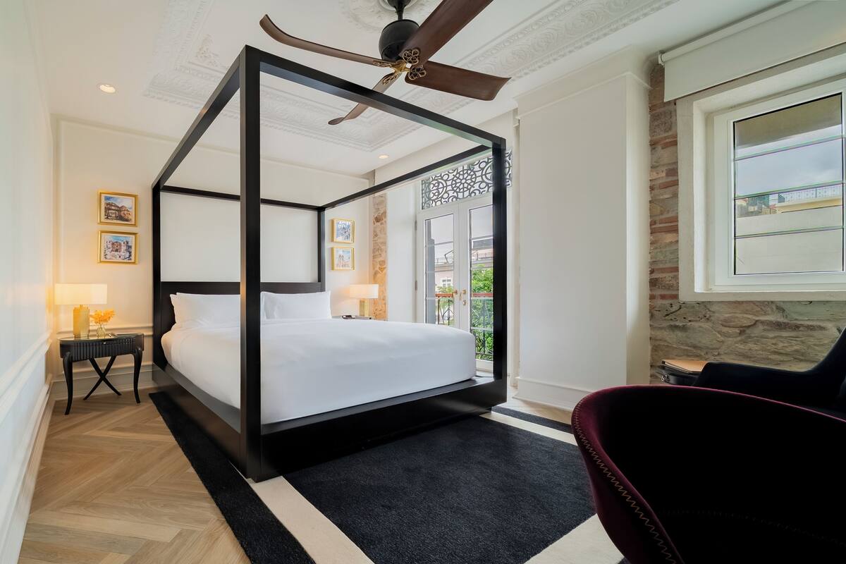 A four-poster bed with a ceiling fan, a black and white rug, a red chair, and a window with a view of the outside.