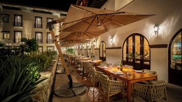 Un espacio exterior para comer con mesas y sillas, sombrillas y un muro de piedra con plantas.
