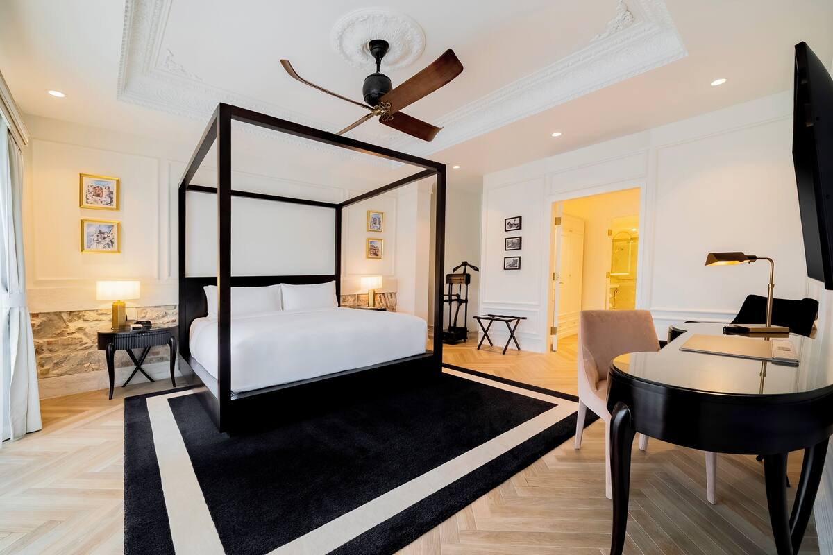 A four-poster bed, a ceiling fan, a desk with a chair, and framed pictures on the walls.