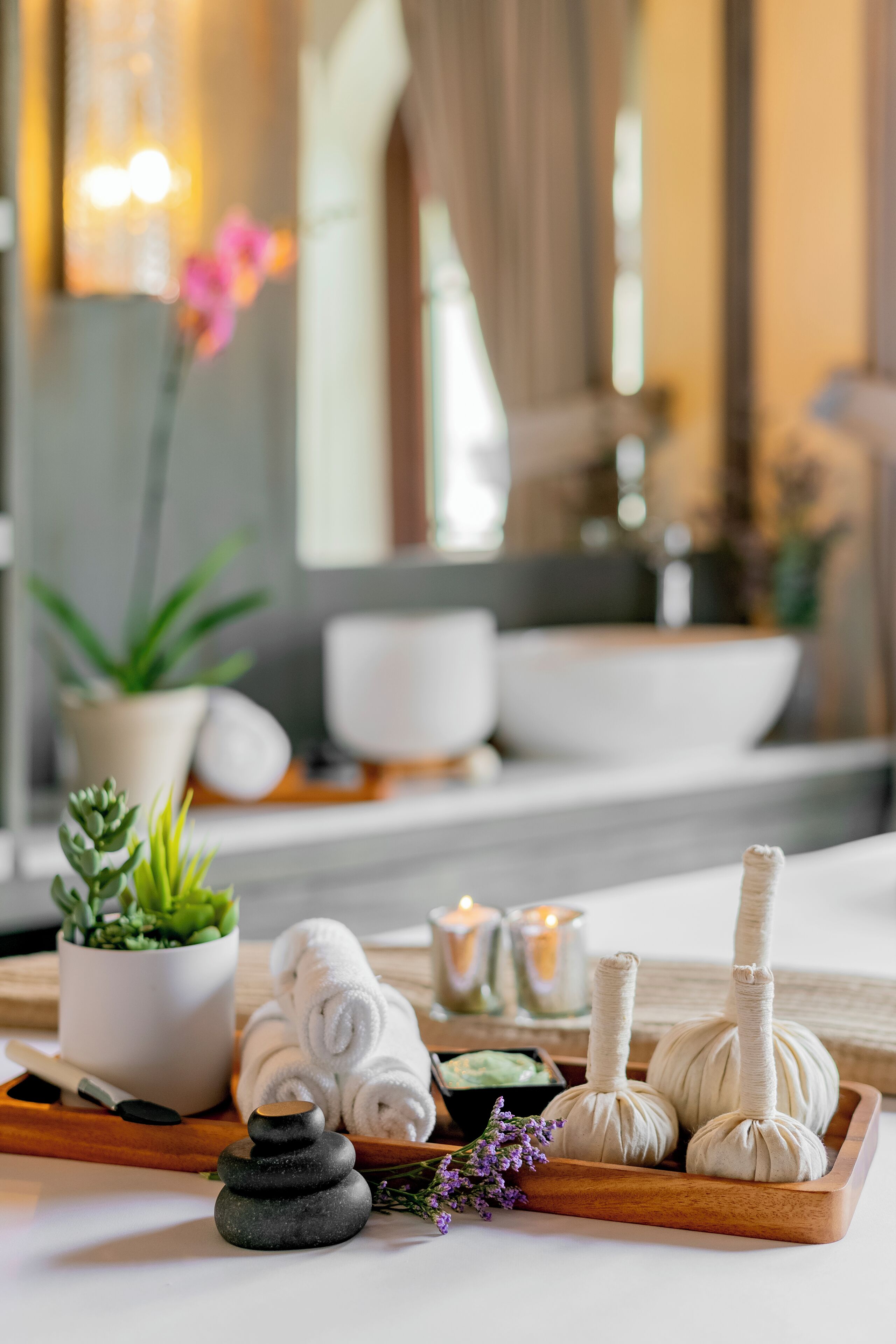 Wellness-Ambiente mit einem Holftablett, das Handtücher, eine kleine Topfpflanze, Kerzen und ein Steinmassagegerät enthält.