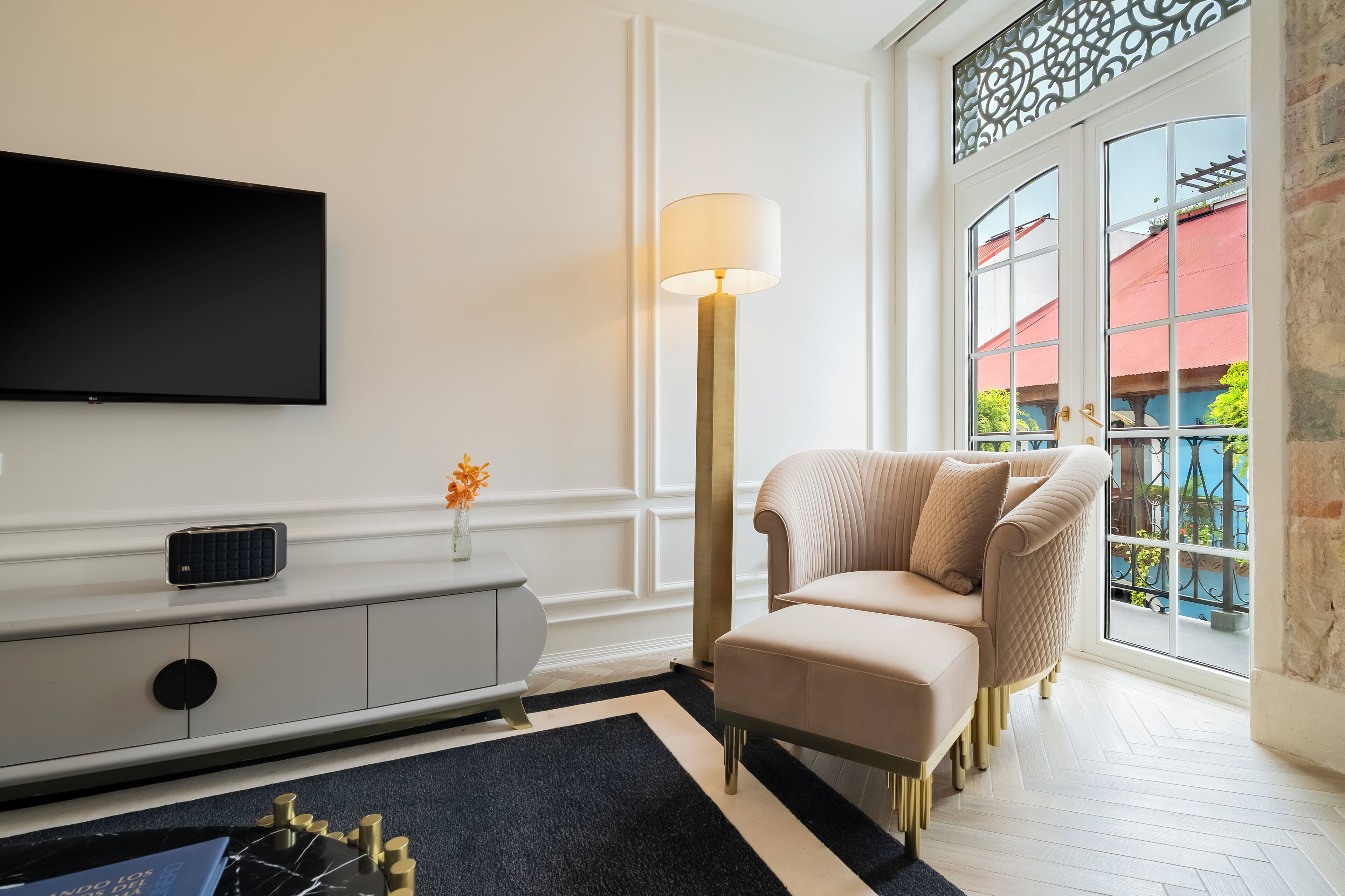 Una sala de estar moderna con televisor de pantalla plana, una lámpara de pie, un sofá color beige y vistas a un edificio de techo rojo a través de un ventanal grande.