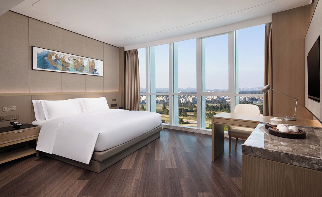 Ein Hotelzimmer mit einem großen Bett, einem Schreibtisch, einem Sessel, einem Fernseher und einem Fenster mit Blick auf die Stadt.