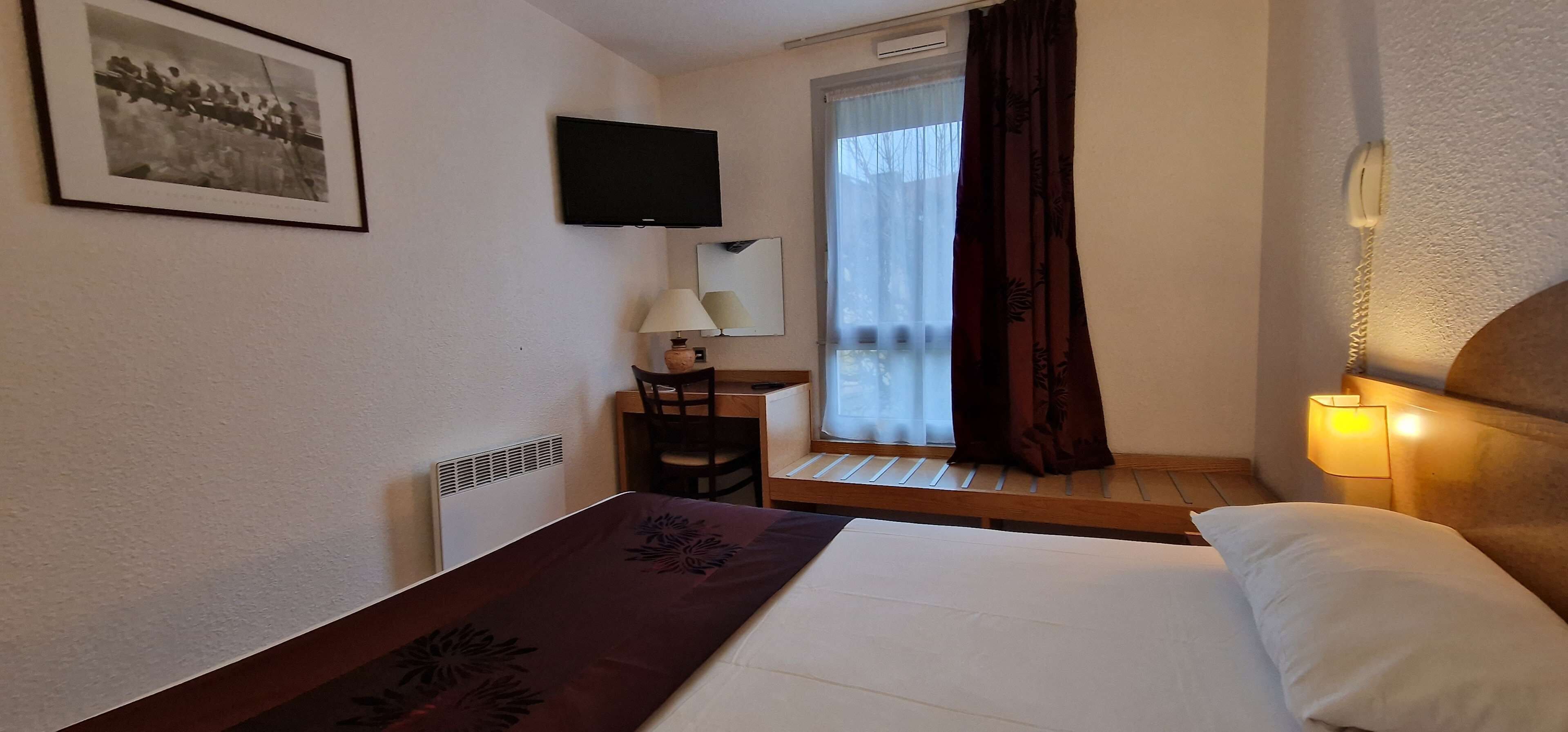 A hotel room with a bed, a desk with a chair, a television, and a window with curtains.