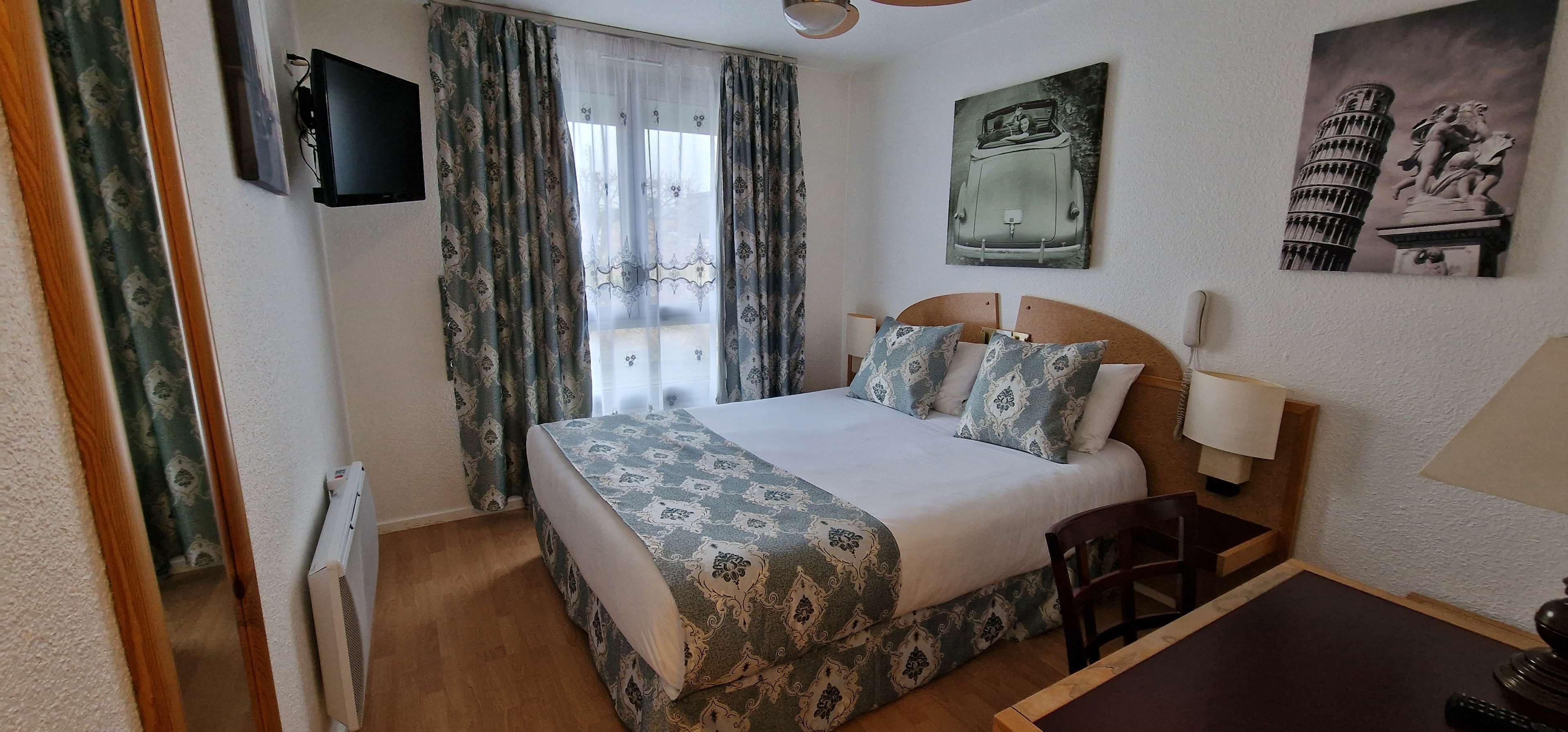 A hotel room with a bed, a desk, a chair, a TV, and artwork on the walls.