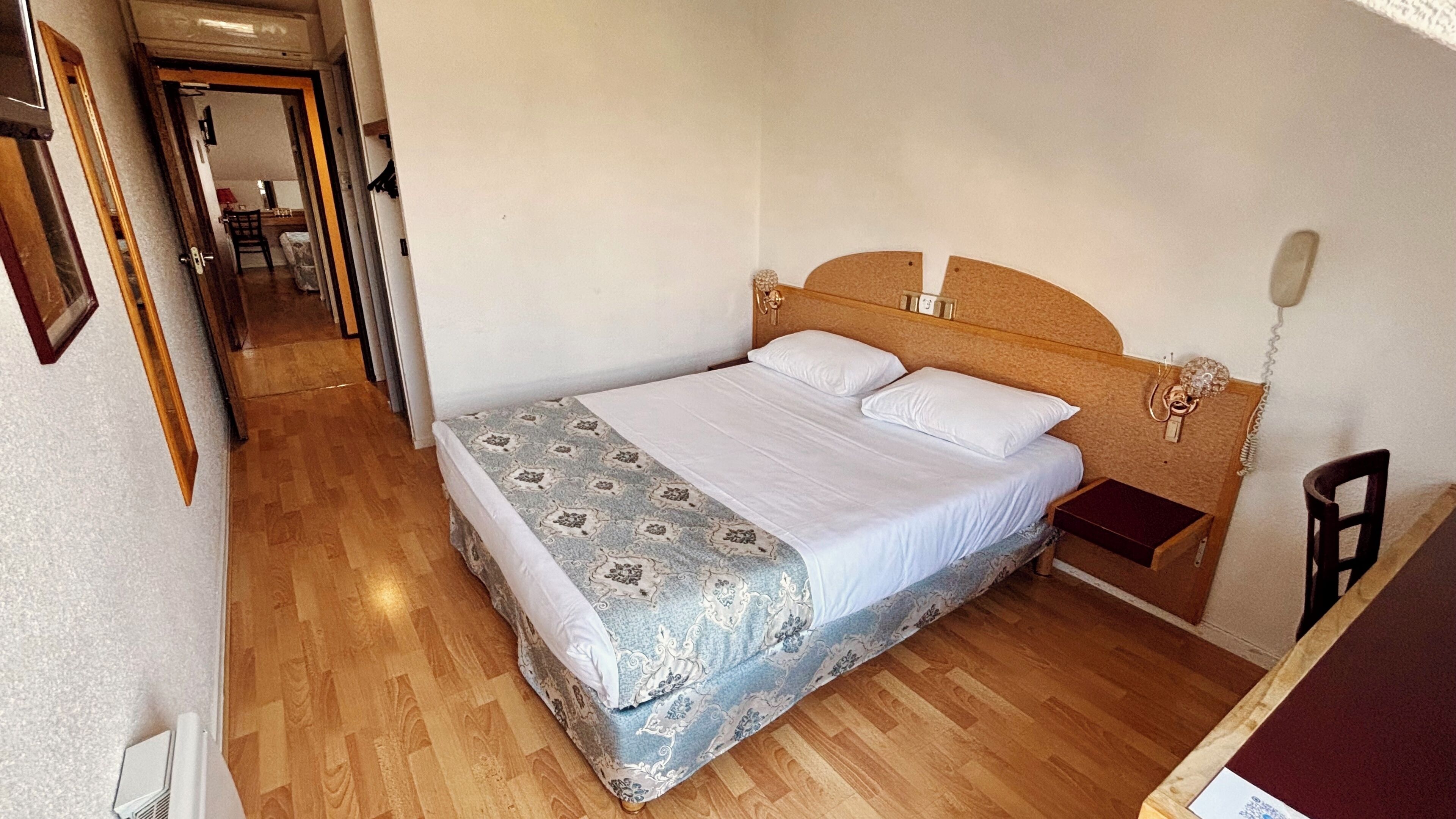 A bedroom with a wooden headboard, a bed with a patterned bedspread, a bedside table, and a chair.