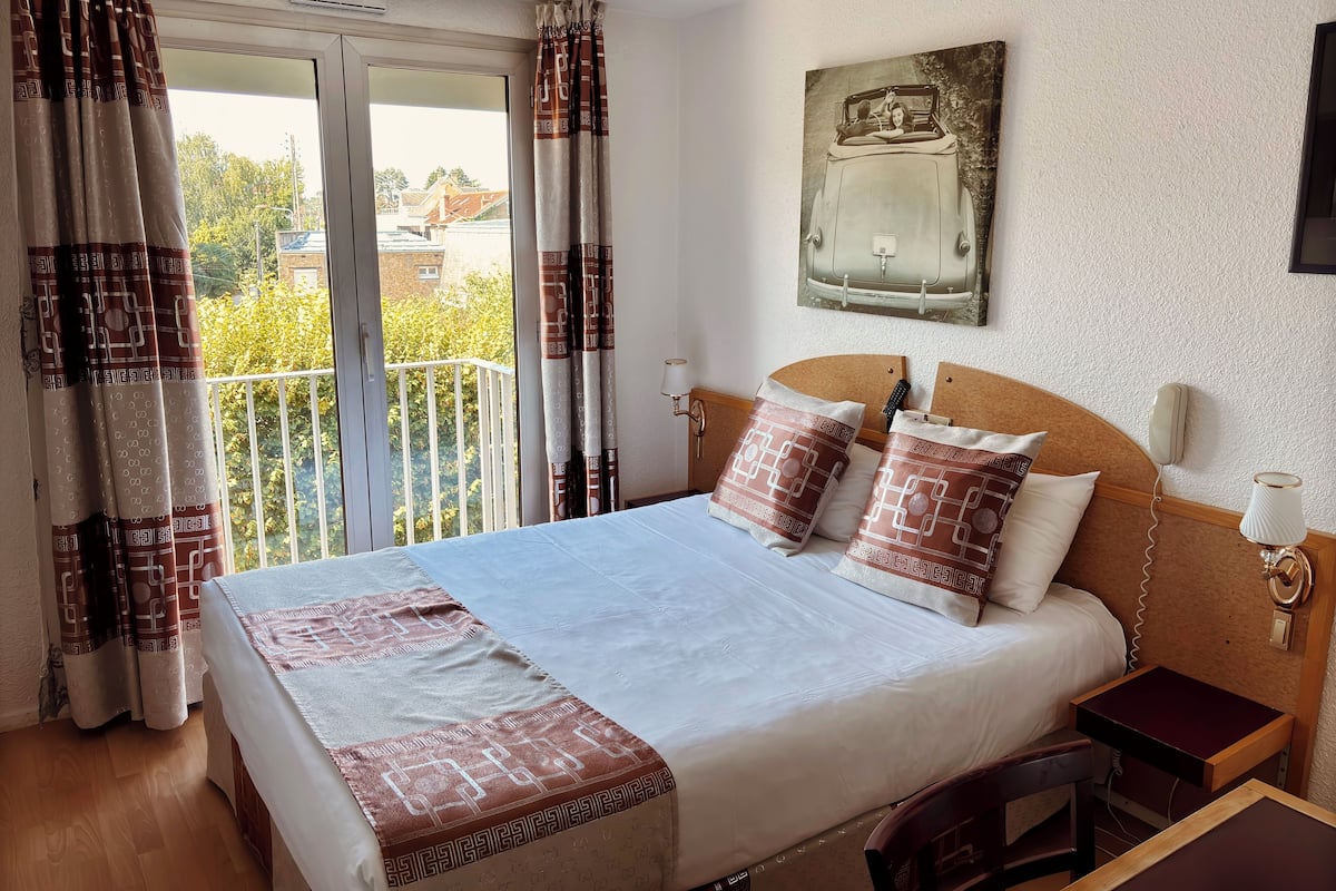 A hotel room with a bed, a bedside table, a chair, a balcony with curtains, and a wall-mounted telephone.