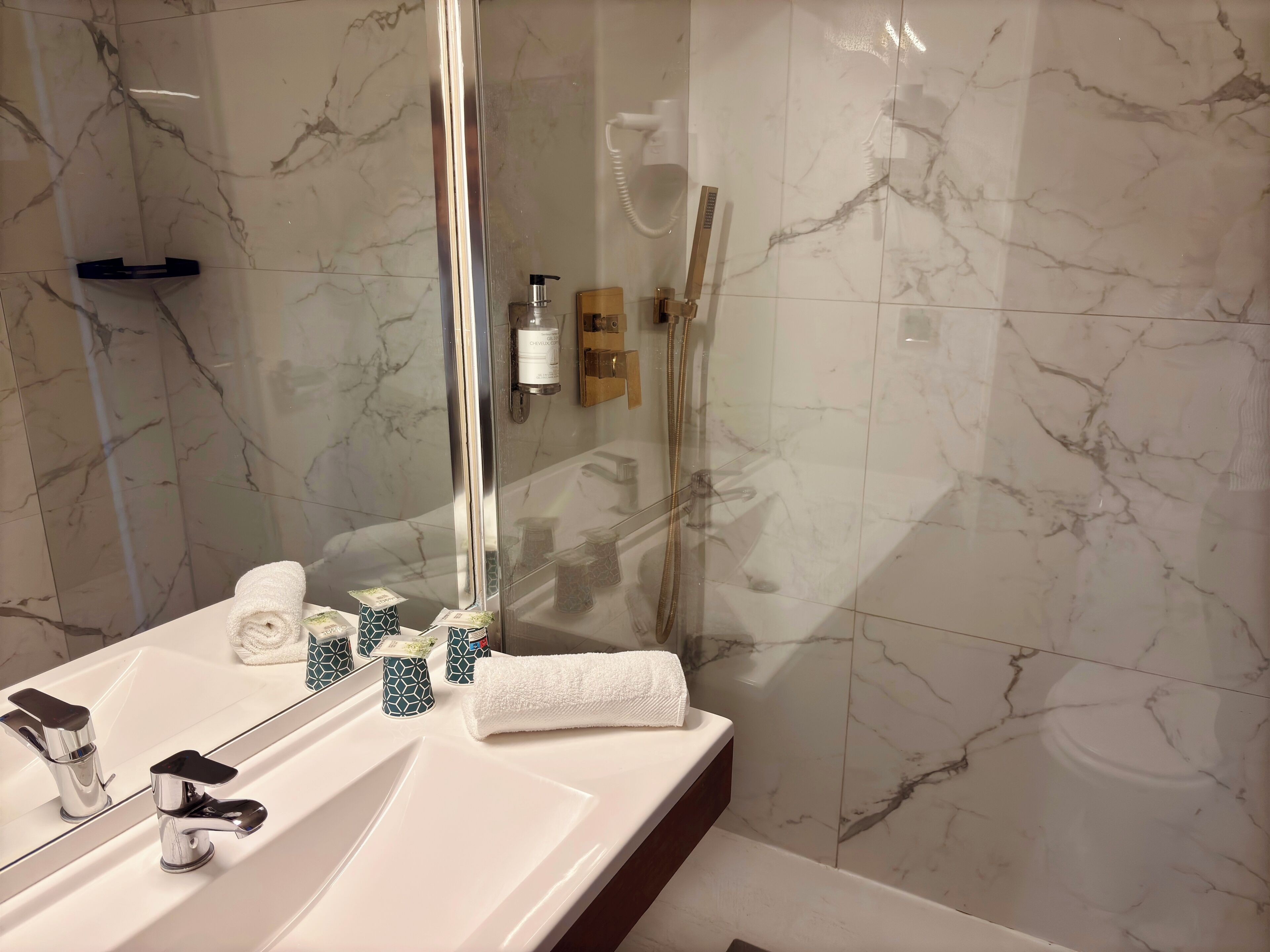 Un baño con pared de azulejos de mármol, un lavamanos blanco con grifería cromada, ducha con cerramiento de vidrio y una toalla sobre la encimera.
