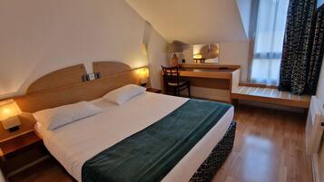 A hotel room with a bed, a desk, a chair, a lamp, and a window with curtains.