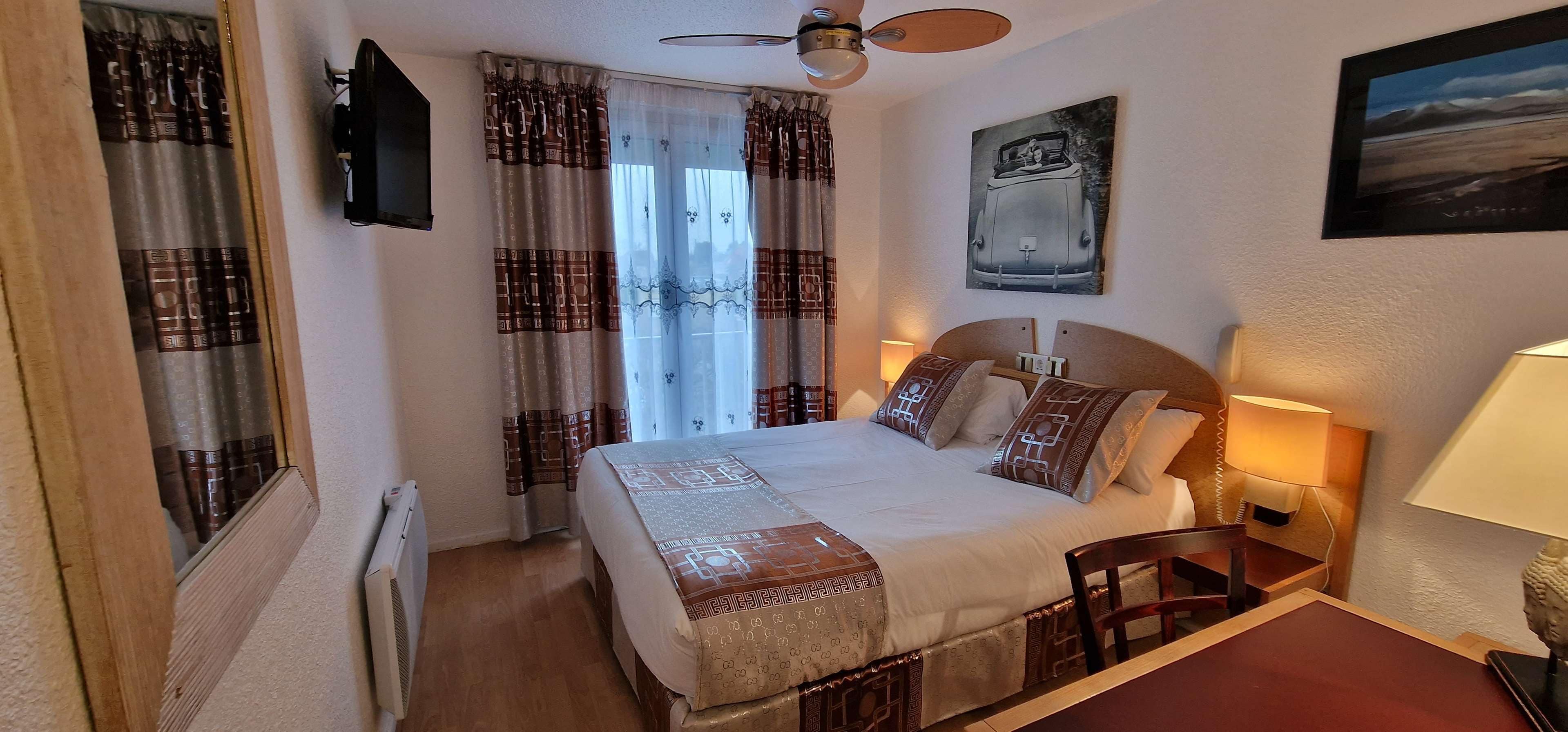 A hotel room with a bed, a ceiling fan, a window with curtains, a wall-mounted TV, and a painting.