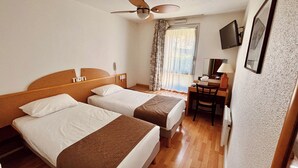 A hotel room with two beds, a desk, a chair, a ceiling fan, a TV, and a window with curtains.