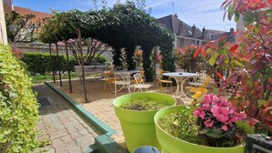 An outdoor patio with tables and chairs, surrounded by greenery and a building in the background.