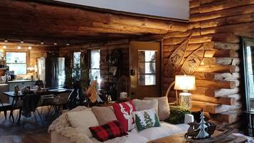 L’intérieur chaleureux d’un chalet en rondins présente un coin repas en bois, un canapé agrémenté de coussins décoratifs et un coffre en bois rustique.