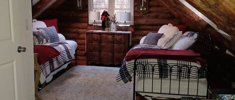 A cozy attic bedroom with two beds, a small desk, and a window with curtains.