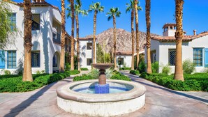 A courtyard with a fountain, palm trees, and buildings with white walls and blue windows.
