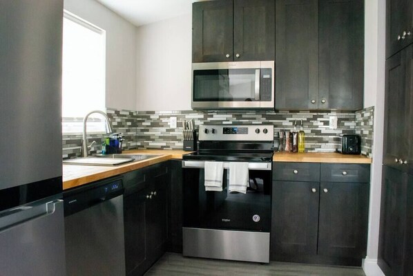 Una cocina moderna con armarios oscuros, lavavajillas de acero inoxidable, microondas, horno y salpicadero de azulejos.