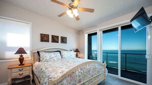 A bedroom with a bed, a nightstand, a ceiling fan, a TV mounted on the wall, and a large window with a view of the ocean. - Promo! Top Floor, Corner Oceanfront Condo - Indoor Pool/Hot Tub/Wifi and HDTV! (Depoe Bay)